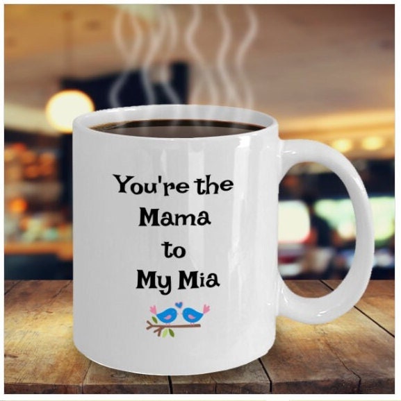 White mug with "You're the Mama to My Mia" text and two blue birds, sitting on a wooden table in a cozy cafe.