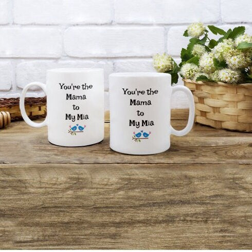 Two white mugs with "You're the Mama to My Mia" text, displayed on a wooden table near flowers and a wicker basket.