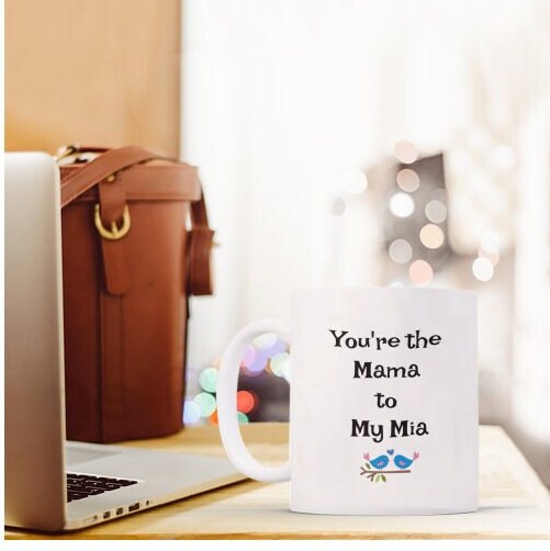 White mug on desk with text "You're the Mama to My Mia," next to a laptop and brown bag, cozy setting.