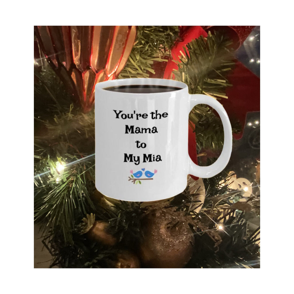White mug with "You're the Mama to My Mia" and bird illustration, displayed in front of Christmas tree decorations.