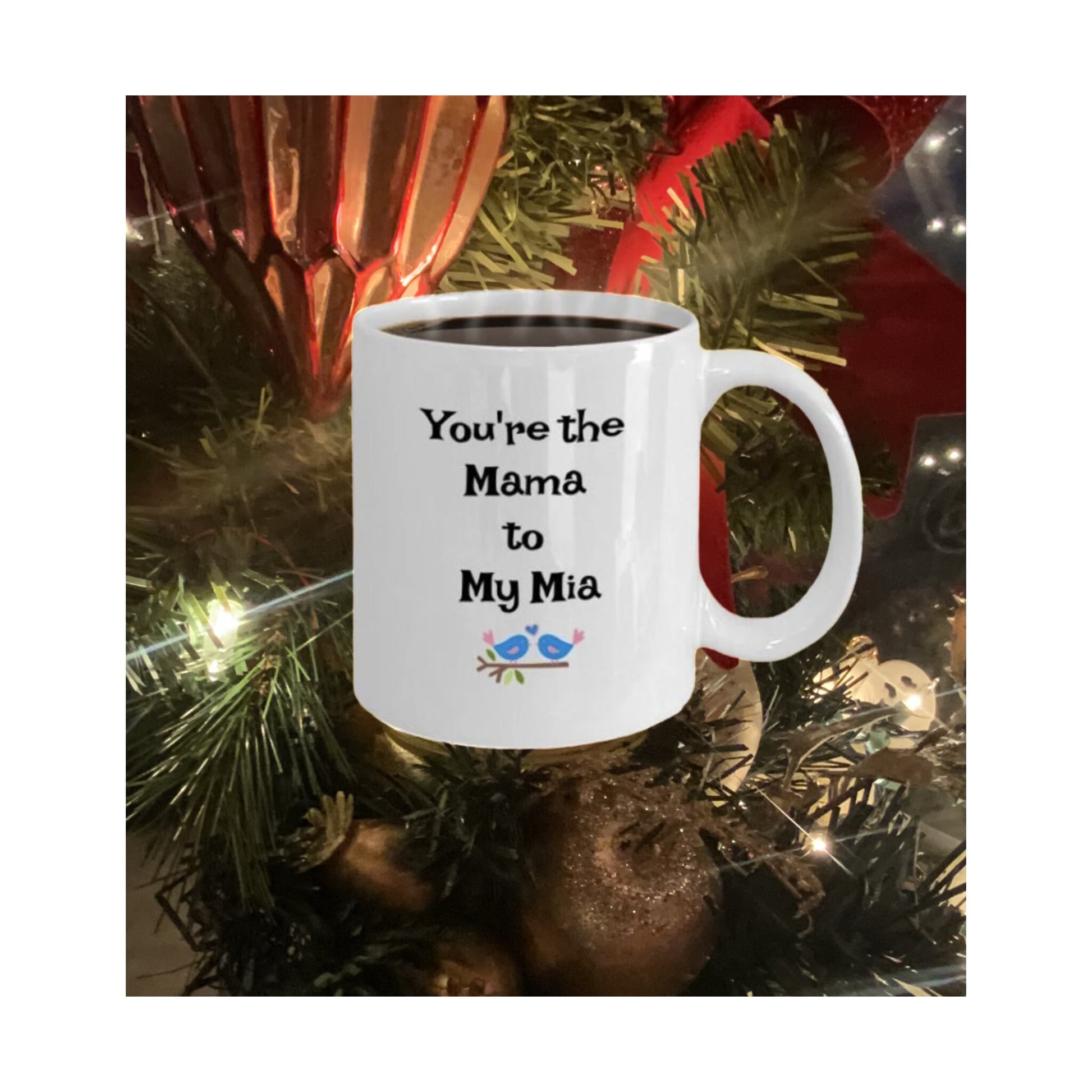 White mug with "You're the Mama to My Mia" and bird illustration, displayed in front of Christmas tree decorations.